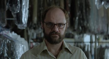 Movie still from “Pity” (2018), directed by Babis Makridis – A man with a goatee wearing glasses and a striped shirt; Close Up shot, Over the shoulder angle