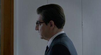 Movie still from “Pity” (2018), directed by Babis Makridis – A person wearing a suit and glasses; Close Up shot, Over the shoulder angle