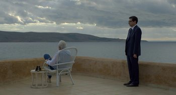 Movie still from “Pity” (2018), directed by Babis Makridis – A man and a woman sitting on a chair by the water; Wide shot, High angle