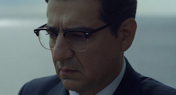 Movie still from “Pity” (2018), directed by Babis Makridis – A person wearing a suit and glasses; Close Up shot, Over the shoulder angle