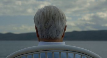 Movie still from “Pity” (2018), directed by Babis Makridis – An older man sitting in a chair looking out at the water; Close Up shot, Low angle