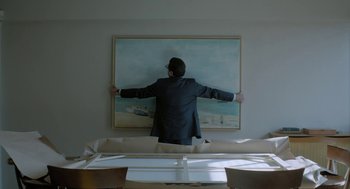 Movie still from “Pity” (2018), directed by Babis Makridis – A man standing on a couch in front of a painting; Medium shot, Low angle