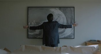 Movie still from “Pity” (2018), directed by Babis Makridis – A man standing in front of a painting of an ocean; Medium shot, Low angle