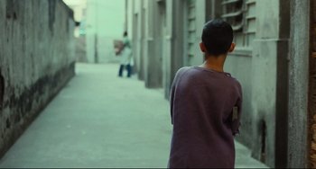 Movie still from “Pixote” (1980), directed by Hector Babenco – A young boy is walking down the street with a cell phone in his hand; Wide shot, Over the shoulder angle