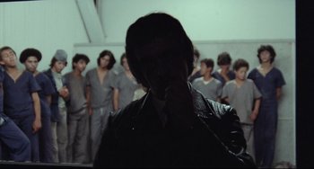 Movie still from “Pixote” (1980), directed by Hector Babenco – A group of people in a dark room with a man in a leather jacket looking at the camera; Medium shot, Over the shoulder angle