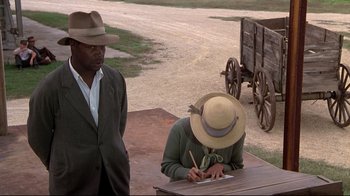 Movie still from “Places in the Heart” (1984), directed by Robert Benton – A man standing next to a woman writing on a paper; Medium shot, Over the shoulder angle