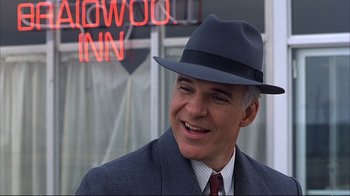 Movie still from “Planes, Trains & Automobiles” (1987), directed by John Hughes – A man wearing a suit and a hat smiling for the camera; Close Up shot, Over the shoulder angle