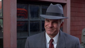 Movie still from “Planes, Trains & Automobiles” (1987), directed by John Hughes – A man in a suit and a hat smiling for the camera; Close Up shot, Over the shoulder angle