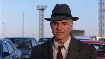 Movie still from “Planes, Trains & Automobiles” (1987), directed by John Hughes – A man wearing a suit and a hat standing in front of some parked cars; Close Up shot, Over the shoulder angle