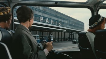 Movie still from “Playtime” (1967), directed by Jacques Tati – A woman is sitting in a car outside of a building; Wide shot, Over the shoulder angle