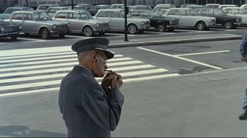 Movie still from “Playtime” (1967), directed by Jacques Tati – A man in a uniform is eating a sandwich; Wide shot, Over the shoulder angle