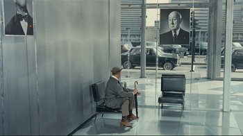 Movie still from “Playtime” (1967), directed by Jacques Tati – An old man sitting on a chair in a lobby; Wide shot, Over the shoulder angle