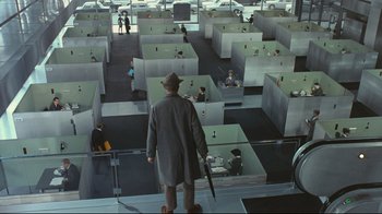 Movie still from “Playtime” (1967), directed by Jacques Tati – A man in a hat and coat holding an umbrella in a room with many cubicles; Wide shot, High angle