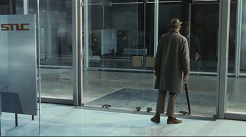Movie still from “Playtime” (1967), directed by Jacques Tati – An older man in a hat and coat is standing in front of a glass door; Wide shot, High angle