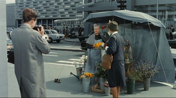 Movie still from “Playtime” (1967), directed by Jacques Tati – A woman is handing flowers to an older woman; Wide shot, Over the shoulder angle
