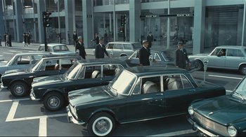 Movie still from “Playtime” (1967), directed by Jacques Tati – A group of men standing next to a line of parked cars; Wide shot, High angle