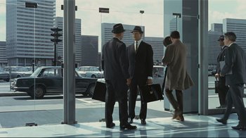 Movie still from “Playtime” (1967), directed by Jacques Tati – A group of men standing next to each other holding suitcases; Wide shot, Over the shoulder angle