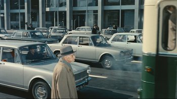Movie still from “Playtime” (1967), directed by Jacques Tati – An older man in a hat and coat walking in a parking lot with parked cars; Wide shot, High angle