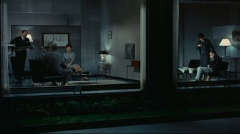 Movie still from “Playtime” (1967), directed by Jacques Tati – Two people sitting on a bench in a living room; Wide shot, Over the shoulder angle