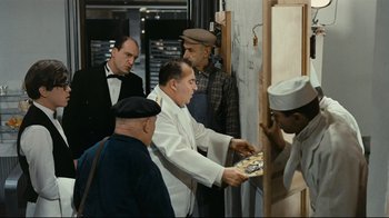 Movie still from “Playtime” (1967), directed by Jacques Tati – A group of men standing next to each other in front of an easel; Medium shot, Over the shoulder angle