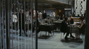 Movie still from “Playtime” (1967), directed by Jacques Tati – A group of people standing around a table with wine glasses on top of it; Wide shot, High angle