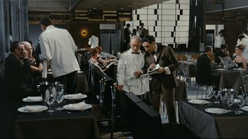 Movie still from “Playtime” (1967), directed by Jacques Tati – Two men in suits are standing in a restaurant; Wide shot, High angle