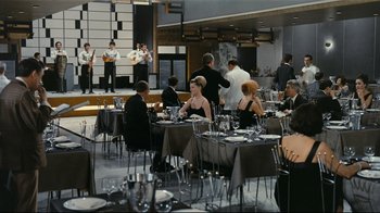 Movie still from “Playtime” (1967), directed by Jacques Tati – A group of people sitting at tables in a room; Wide shot, High angle