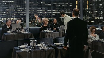 Movie still from “Playtime” (1967), directed by Jacques Tati – A group of people sitting at a table in front of a window; Wide shot, Over the shoulder angle