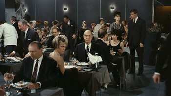 Movie still from “Playtime” (1967), directed by Jacques Tati – A group of people sitting at a table in a room; Wide shot, High angle