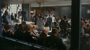 Movie still from “Playtime” (1967), directed by Jacques Tati – A group of people standing around a room; Extreme Wide shot, High angle