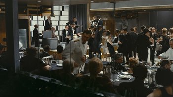 Movie still from “Playtime” (1967), directed by Jacques Tati – A group of people sitting at tables in a room; Wide shot, High angle