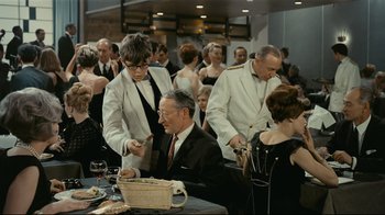 Movie still from “Playtime” (1967), directed by Jacques Tati – A group of people standing around a table with food on top of it; Medium shot, High angle