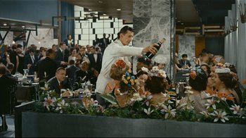 Movie still from “Playtime” (1967), directed by Jacques Tati – A man pouring champagne into a glass bottle at an event; Wide shot, Over the shoulder angle