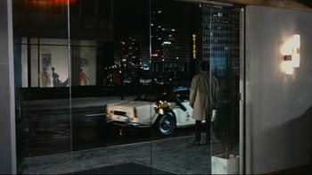 Movie still from “Playtime” (1967), directed by Jacques Tati – A man standing in front of a car on the street; Wide shot, Over the shoulder angle