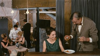 Movie still from “Playtime” (1967), directed by Jacques Tati – A woman sitting at a table in front of a plate of food; Medium shot, Over the shoulder angle