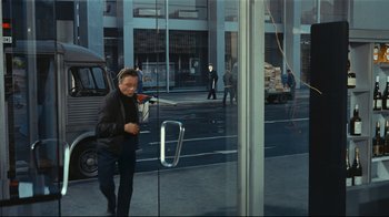 Movie still from “Playtime” (1967), directed by Jacques Tati – A man walking in front of a glass door on a street; Wide shot, Over the shoulder angle
