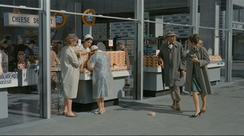 Movie still from “Playtime” (1967), directed by Jacques Tati – A group of people standing around a table with food on top of it; Wide shot, High angle