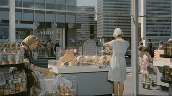 Movie still from “Playtime” (1967), directed by Jacques Tati – A woman standing in front of a cart of food; Wide shot, Over the shoulder angle