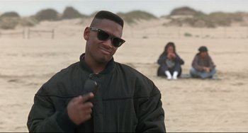 Movie still from “Poetic Justice” (1993), directed by John Singleton – A man in a black jacket holding a cell phone on the beach; Medium shot, Over the shoulder angle