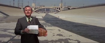 Movie still from “Point Blank” (1967), directed by John Boorman – A man in a suit and tie holding a package; Wide shot, Low angle