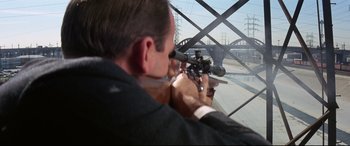Movie still from “Point Blank” (1967), directed by John Boorman – A man in a suit and tie holding a rifle; Medium shot, Over the shoulder angle