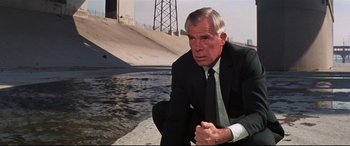 Movie still from “Point Blank” (1967), directed by John Boorman – A man in a suit and tie sitting in front of a body of water; Medium shot, Low angle