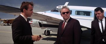 Movie still from “Point Blank” (1967), directed by John Boorman – A man in a suit and tie standing next to a plane; Medium shot, Low angle