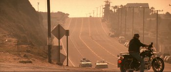 Movie still from “Point Break” (1991), directed by Kathryn Bigelow – Cars are driving down a dusty road on a hazy day; Extreme Wide shot, Low angle