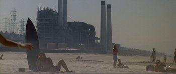 Movie still from “Point Break” (1991), directed by Kathryn Bigelow – A group of people sitting on top of a sandy beach next to a factory; Extreme Wide shot, High angle