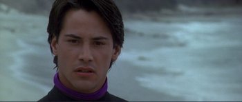 Movie still from “Point Break” (1991), directed by Kathryn Bigelow – A man with long dark hair wearing a black and purple shirt; Close Up shot, Over the shoulder angle