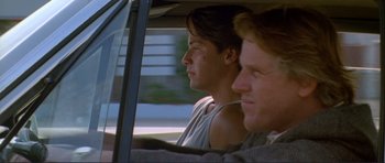 Movie still from “Point Break” (1991), directed by Kathryn Bigelow – Two men are driving in a car and one is looking at the camera; Close Up shot, Over the shoulder angle