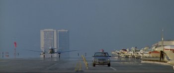 Movie still from “Point Break” (1991), directed by Kathryn Bigelow – An airplane is on a runway with a car on the road; Extreme Wide shot, Low angle