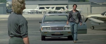 Movie still from “Point Break” (1991), directed by Kathryn Bigelow – A man standing in front of a car in a parking lot; Wide shot, Low angle
