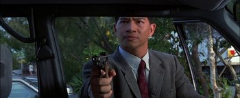 Movie still from “First Strike” (1996), directed by Stanley Tong – A man in a suit and tie holding a gun in his hand; Close Up shot, Over the shoulder angle